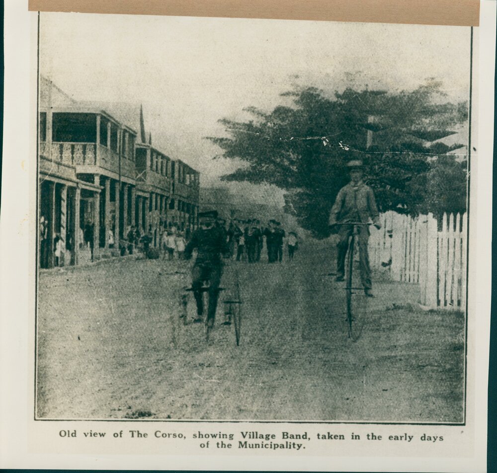 A portrait of two men on bicycles, with the old view of The Corso, with the  Manly Village Band