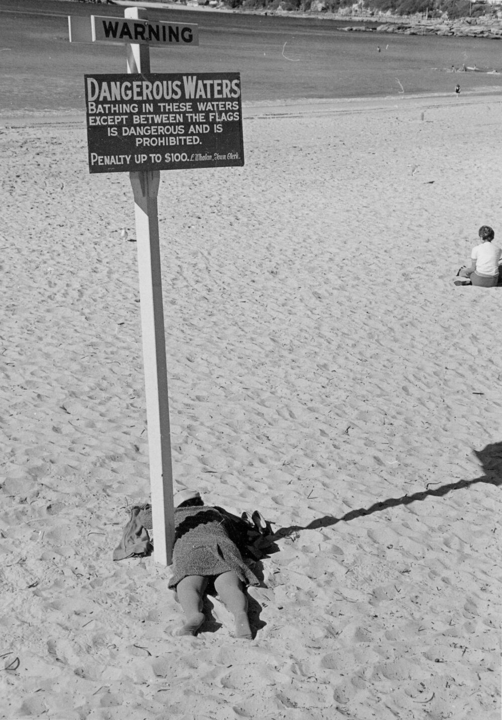 Person laying face-down under a warning sign at Manly Beach, Manly, 1967-1973