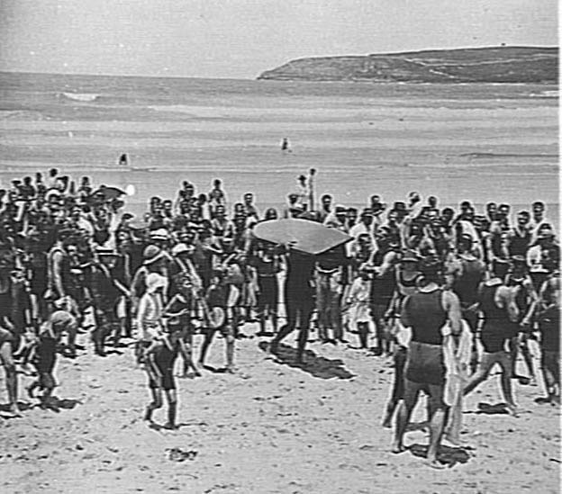 Crowd watching Duke Kahanamoku carrying his surfboard, c 1915
