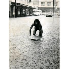 Surfboard rider in flooded Central Avenue, Manly 1970s