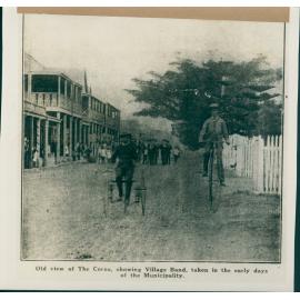 A portrait of two men on bicycles, with the old view of The Corso, with the  Manly Village Band