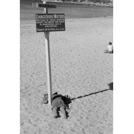 Person laying face-down under a warning sign at Manly Beach, Manly, 1967-1973